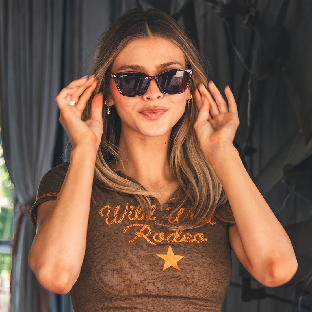 A female model wearing Oneday Fallon sunglasses in Tort Brown, posing with her hands near her sunglasses.