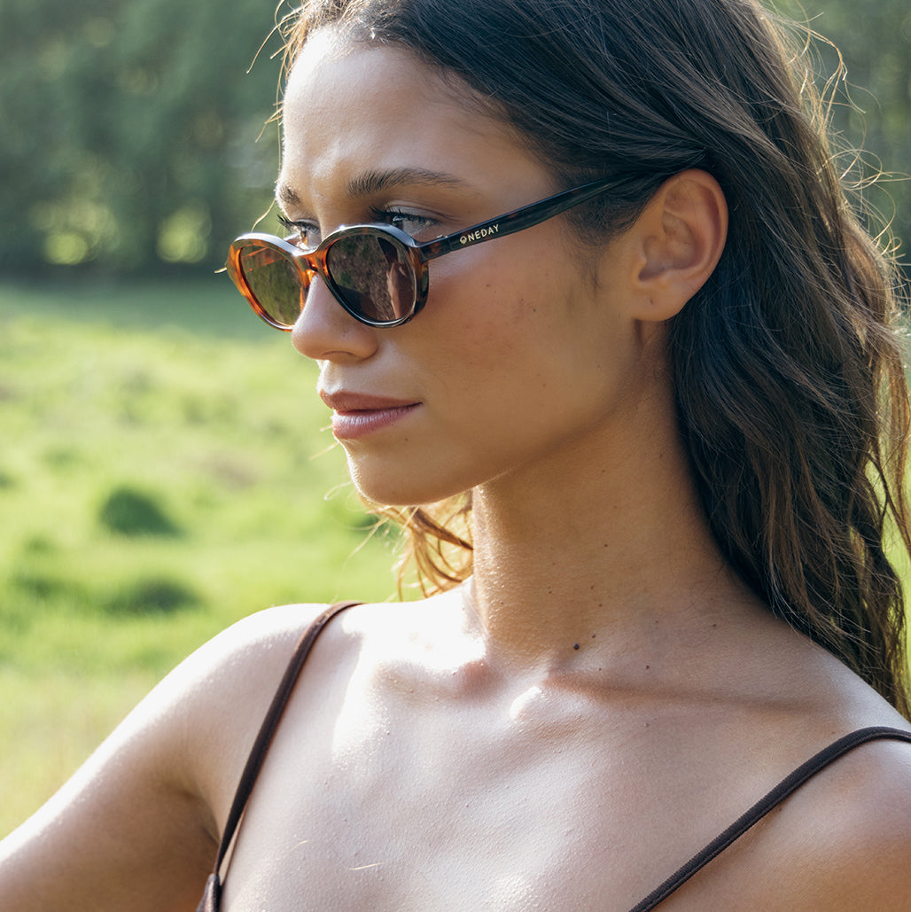 A woman in profile wearing tortoiseshell Oneday Australia's Montana sunglasses with brown lenses, photographed outdoors against a blurred green grassy background. 