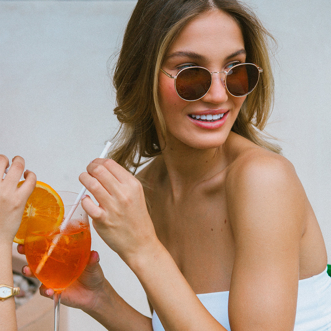 The Inner Circle Gold Round Womens Sunglasses made of metal and brown lens on model drinking an orange drink